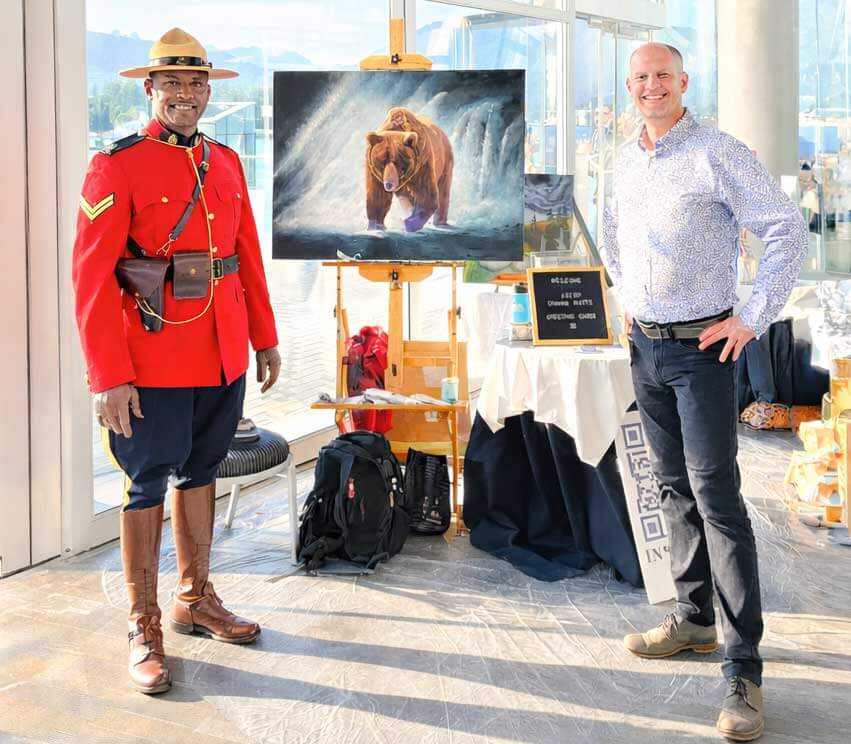 featured artist graham watts and canadian mountie one in a red uniform, standing in front of his oil painting of a bear.  