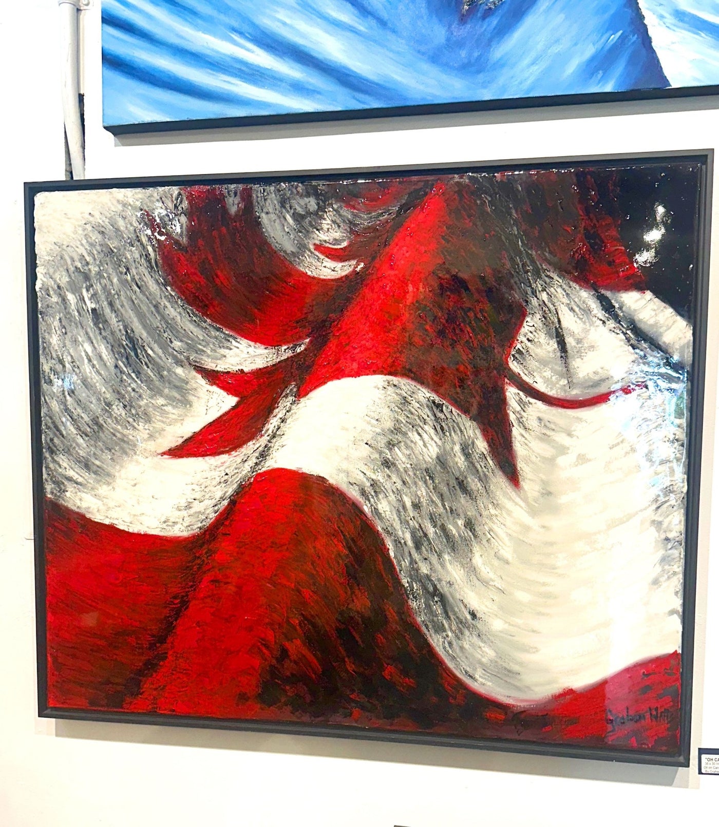 An abstract canvas painting with bold red, white, and black brushstrokes, housed in a black frame and displayed on a gallery wall below another blue-toned painting.